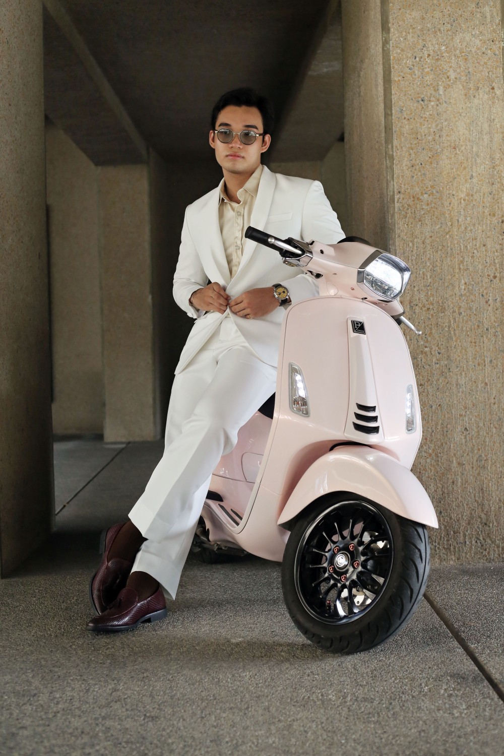 Man in a tailored white suit with beige shirt, leaning on a pink scooter in an urban setting