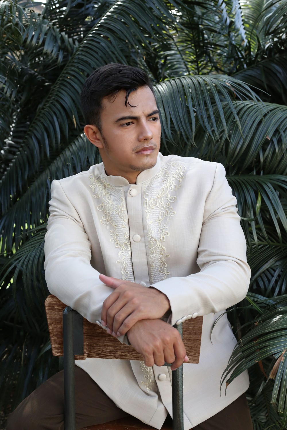 Man wearing a cream piña silk Barong Tagalog with embroidered details, seated outdoors with tropical foliage background